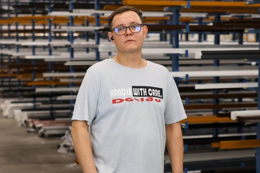 Kamil Rejdak Mann im Lager in weißem T-Shirt mit der Aufschrift 'Handle with care' und Brille.